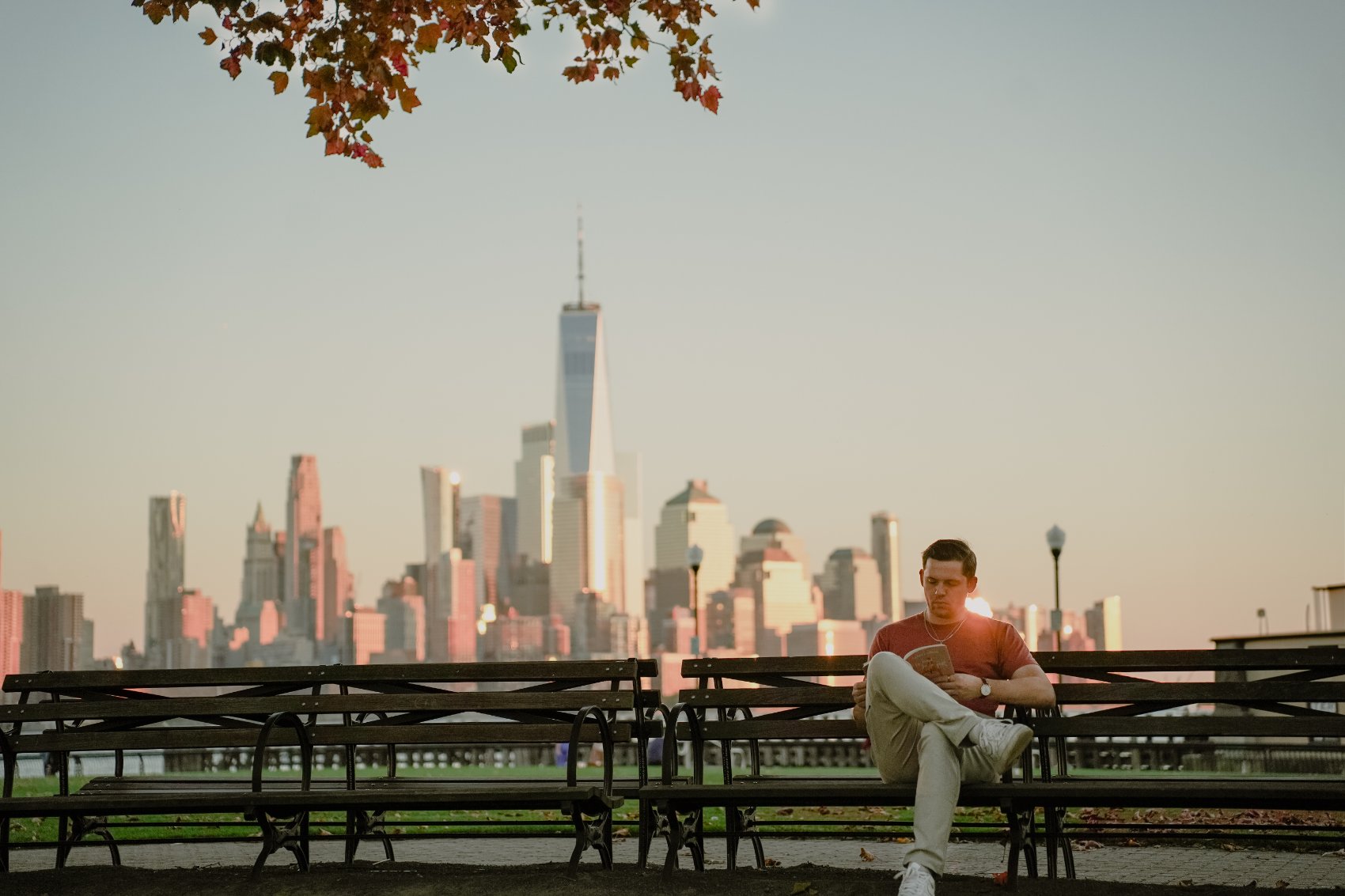 Michael reading on a park bench with the New York City skyline at sunset